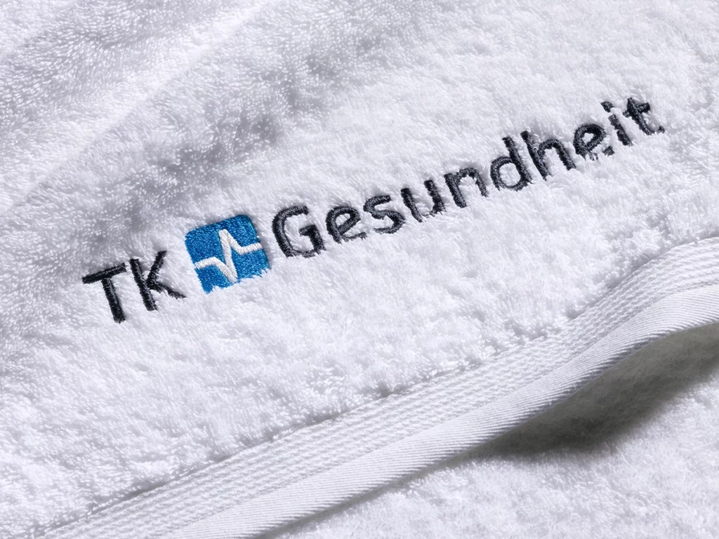 Close up auf weißes Frottierhandtuch mit zweifarbig gesticktem TK Gesundheit Logo. Flauschiger Frottier mit Biesenbordüre von MÖVE Professional