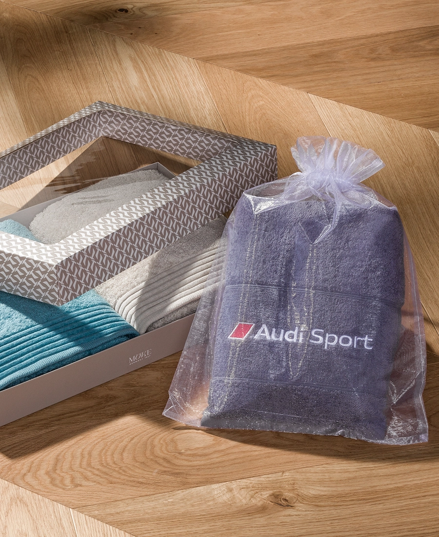 Zwei gefaltete LOFT-Handtücher in einer halb geöffneten Geschenkbox und ein mit Audi Sport besticktes Handtuch im transparenten Organzabeutel