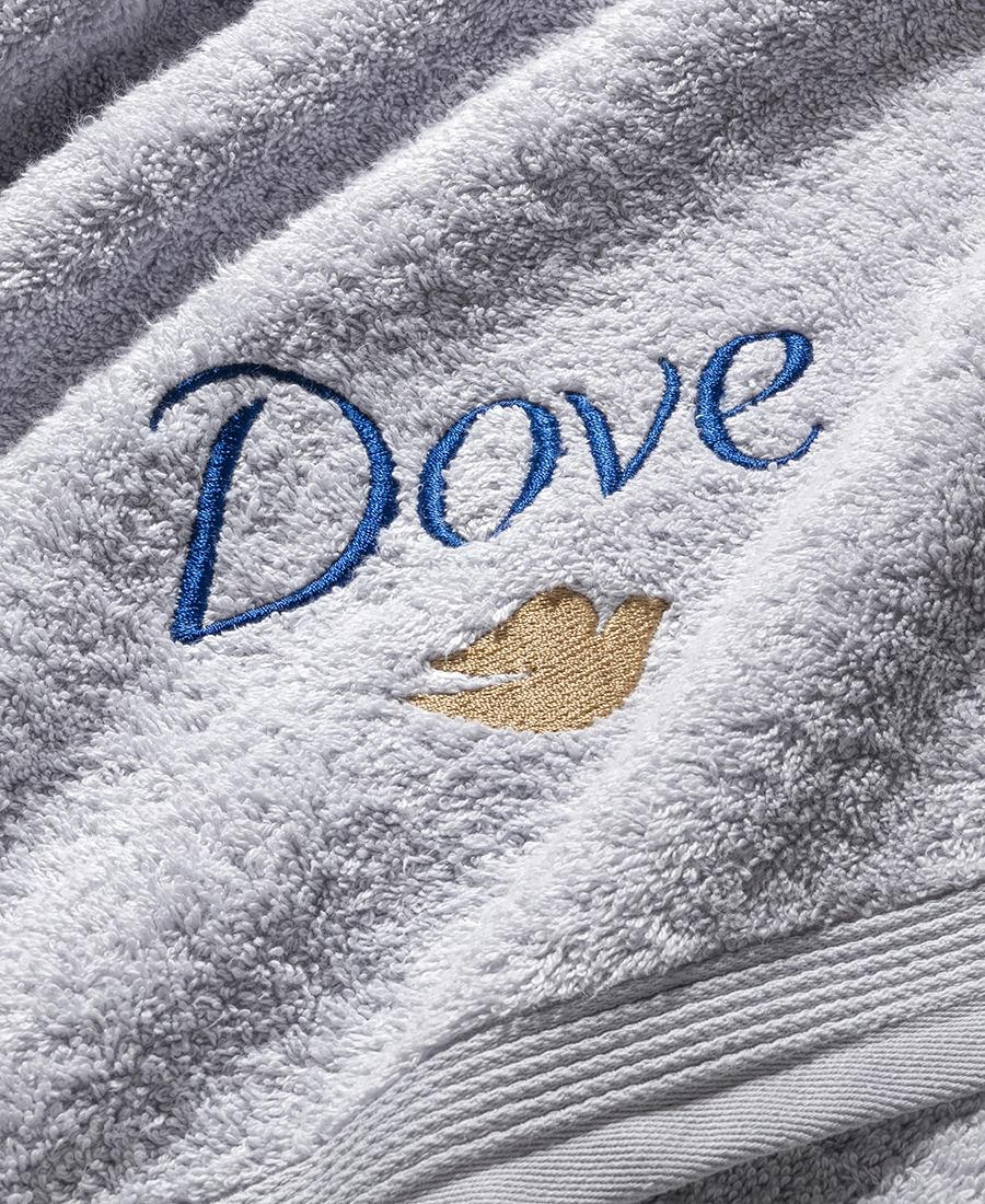 Nahaufnahme eines zweifarbig gestickten DOVE-Logos auf einem silbergrauen Handtuch mit Biesenbordüre